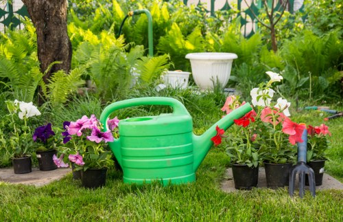A garden scene featuring a neatly maintained grassy area bordered by potted flowering plants, including pink, purple, and white blooms. In the foreground, there is a green plastic watering can positioned on the grass, surrounded by small black pots with vibrant flowering plants. The background shows a variety of lush green foliage, trees, and shrubs, with some garden tools such as a red-handled fork resting on the ground. The overall setting appears to be part of a well-kept outdoor space, reflecting typical garden maintenance activity, suitable for gardening services in Leyton, with natural lighting indicating a bright, partly sunny day with clear weather conditions.